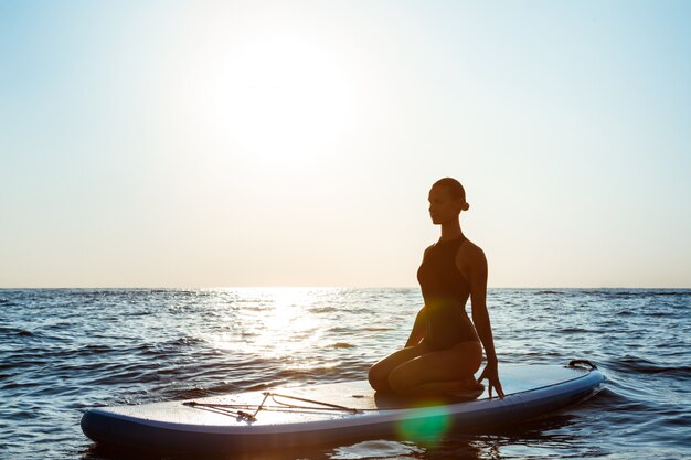 Silhouette di una bella donna che pratica yoga su una tavola da surf all'alba 176420 2034